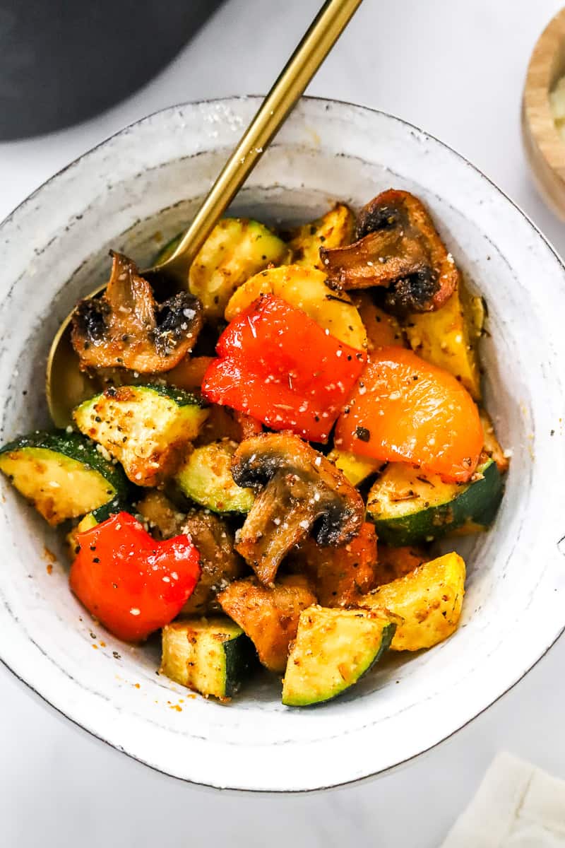 Roasted zucchini, squash, mushrooms and peppers in a round bowl with a spoon in the bowl.